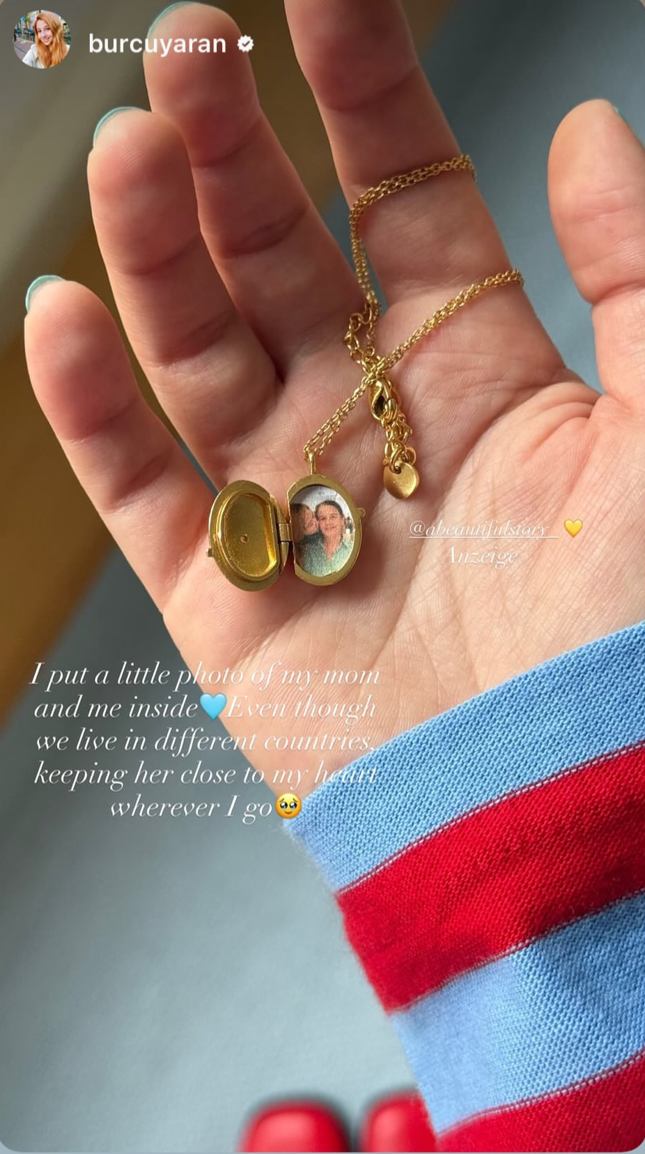 A close-up of a hand wearing a delicate gold chain necklace with two oval charms, one plain gold and the other featuring a colorful floral design. The hand is resting on a gray smartphone screen, wearing a red and blue striped sweater.